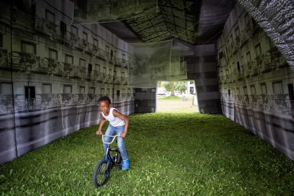 A kid on a bicycle rides through an inflatable structure with images of a balcony apartment on the walls