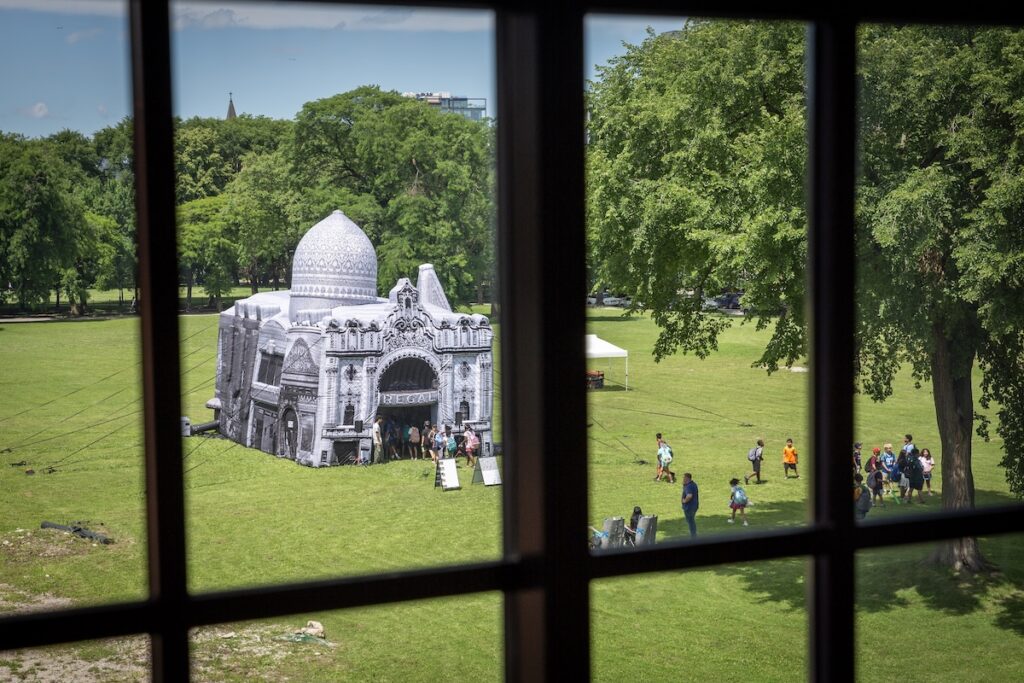 Through a windowpane, an inflatable structure draws crowds in a green field