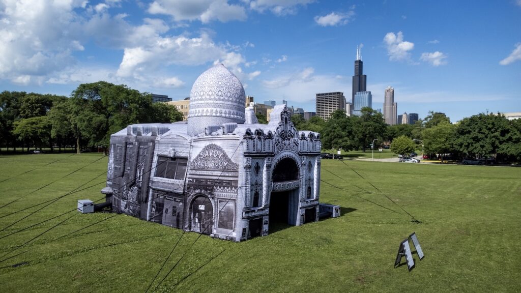 People gather in a field around a white inflatable building with the Chicago skyline in the distance