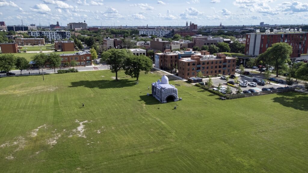 An aerial view of a white inflatable structure in a grassy field, with low rise buildings in the background