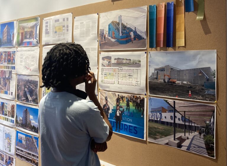 From behind, a person looking at a cork board with pinned up architectural plans and renderings
