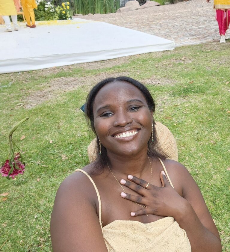 A dark-skinned femme person looking at the camera with a big smile and a hand on her chest. The background is green grass, flowers, and pavement.