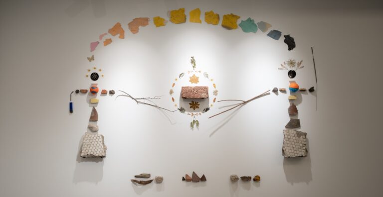 A wall mural made of flowers, leaves, sticks, tiles, paint chips, with a brick at center