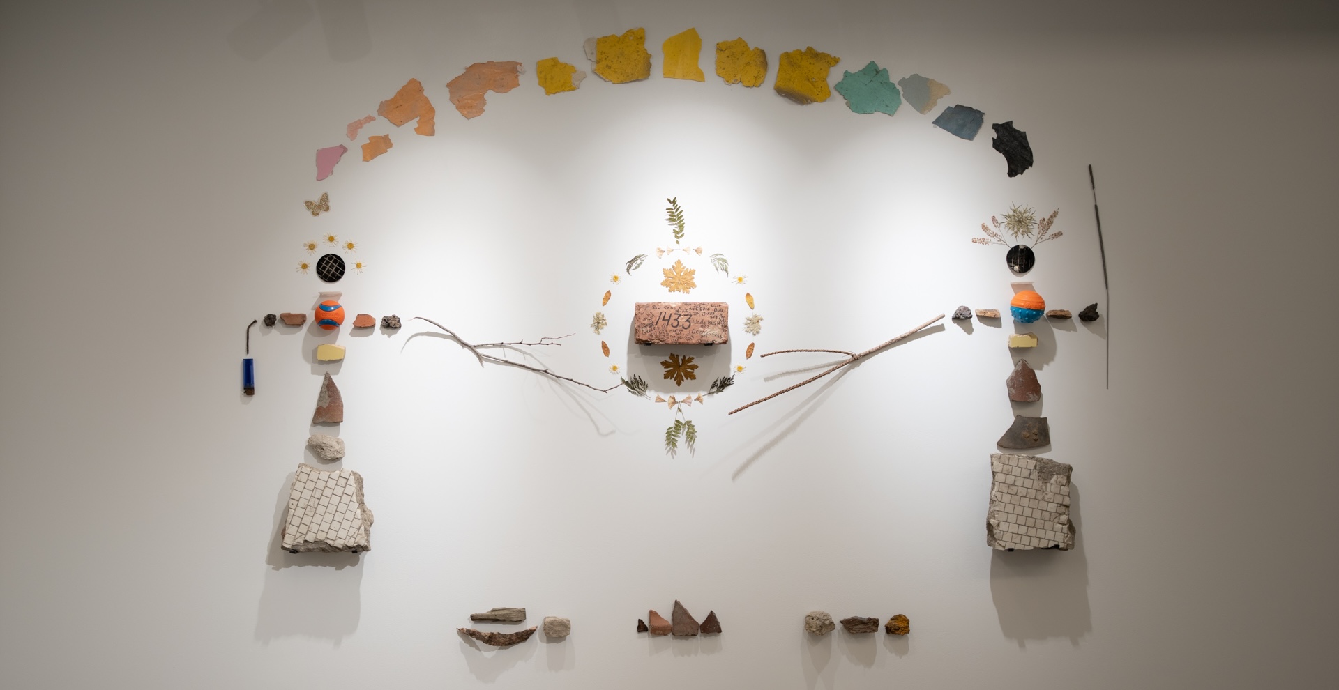 A wall mural made of flowers, leaves, sticks, tiles, paint chips, with a brick at center